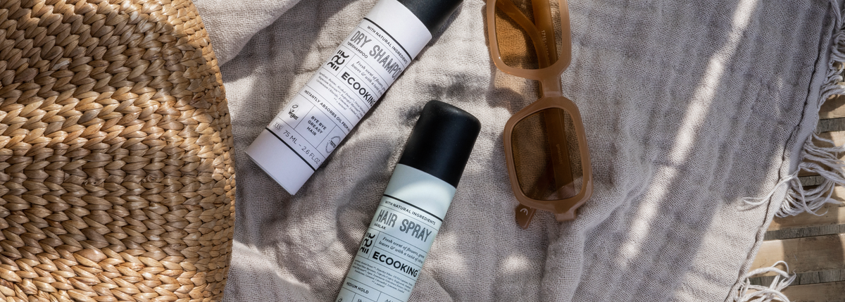 Dry Shampoo og Hair Spray fra ECOOKING liggende på et sandfarvet hørstof sammen med en kurvetaske og et par solbriller.