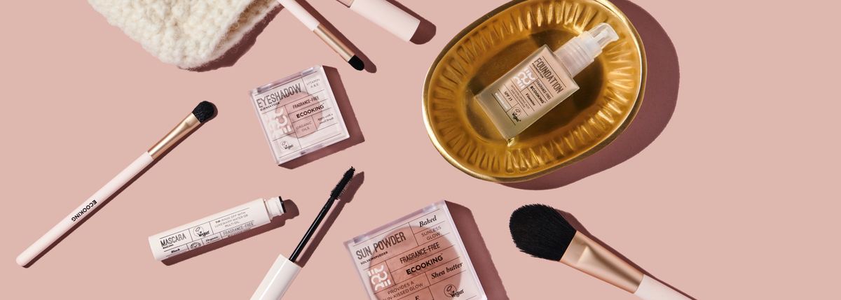 Makeup fra ECOOKING lagt ud på en støvet lyserød baggrund: pensler, øjenskygge, mascara, læbestift, Sun Powder og en foundation i et guldfarvet fad.