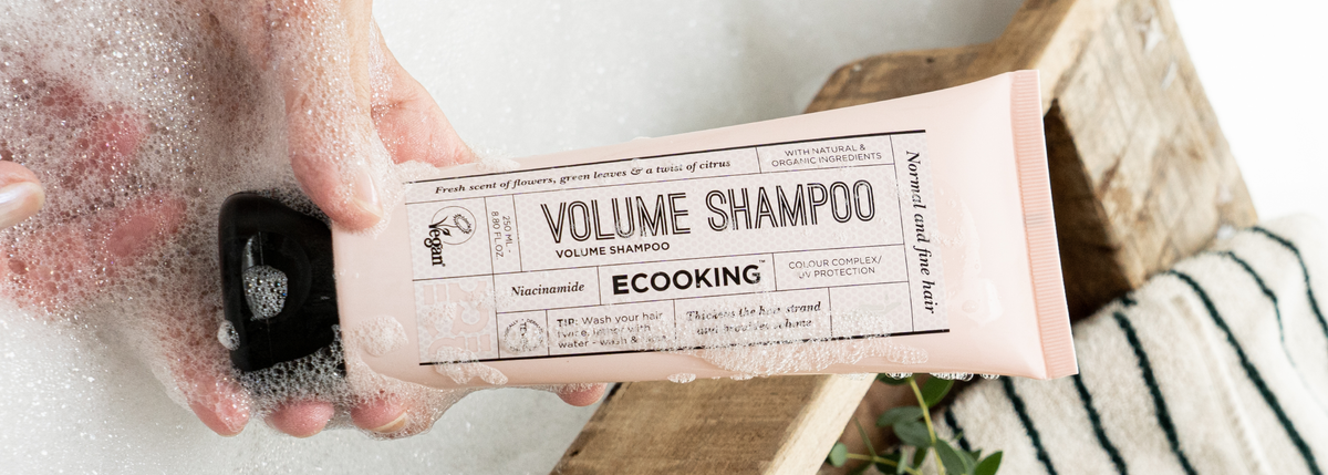 En hånd holder en lyserød tube Volume Shampoo fra ECOOKING over skum i et badekar.
