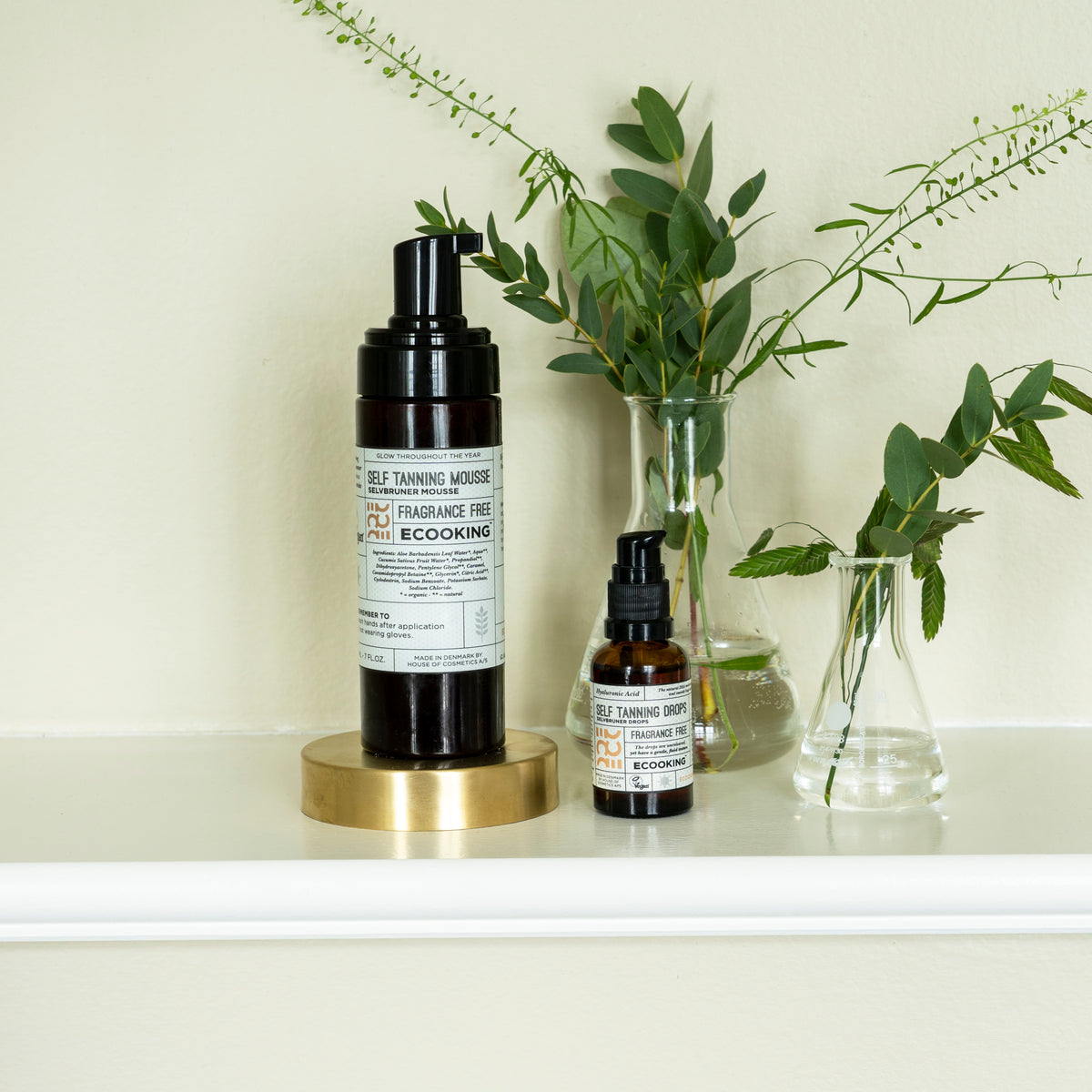 Selvbruner Dråber og Selvbruner Mousse på en hylde med grønne grene i glasvaser – selvbruner dråber og tanning drops til ansigt og krop
