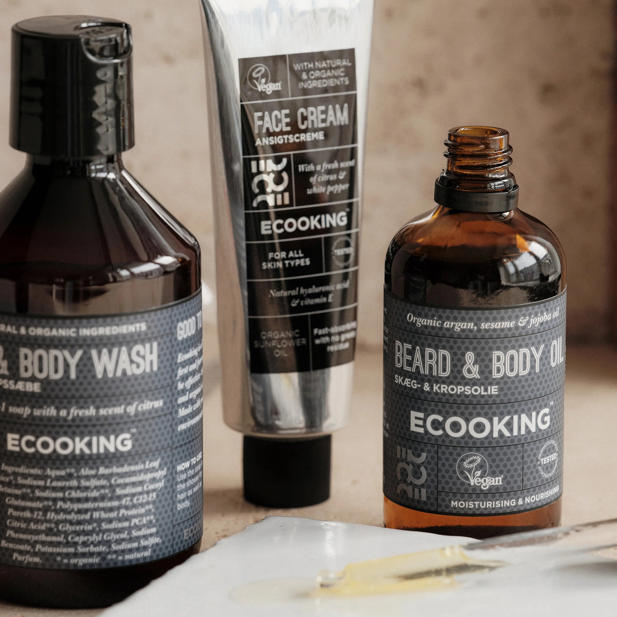 ECOOKING Skæg- og Kropsolie i brun glasflaske sammen med ECOOKING Men Face Cream og Hair & Body Wash på en lys stenoverflade – skægolie og kropsolie til mænd, pleje ved skægpest