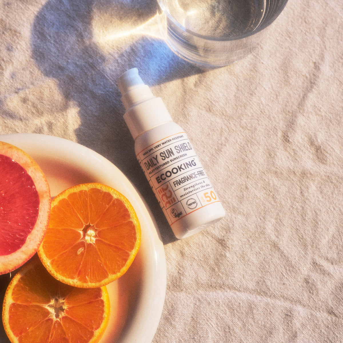 ECOOKING Ultralet Solcreme SPF 50 i sollys ved siden af grapefrugt, appelsinskiver og et glas vand – let solcreme med solfaktor 50, der ikke fedter eller tilstopper porer